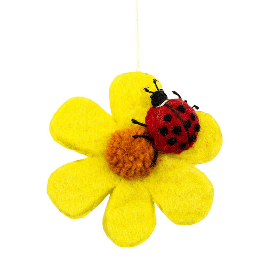 A felt flower ornament with bright yellow petals, an orange-brown center, and a red ladybug with black spots resting on top, hanging by a thin string against a white background.