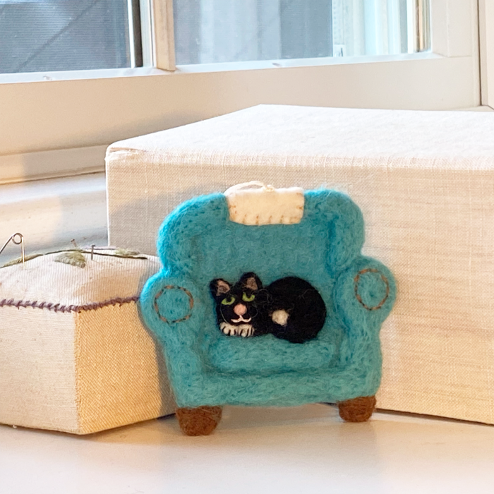 A small felted wool chair in turquoise blue with a black-and-white cat figurine inside, placed on a white surface beside a cream fabric-covered box near a window.