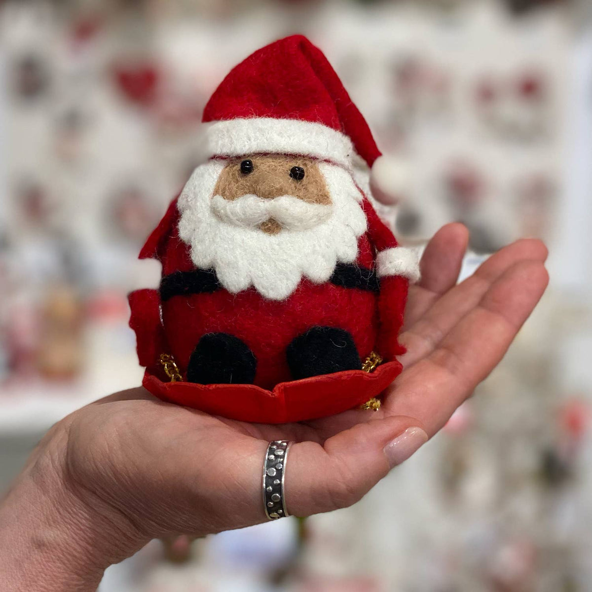 A small felt Santa Claus ornament being held in a person's hand. The ornament is made of red felt with a white beard and trim, black pants, and gold jingle accents, with a softly blurred holiday-themed background.