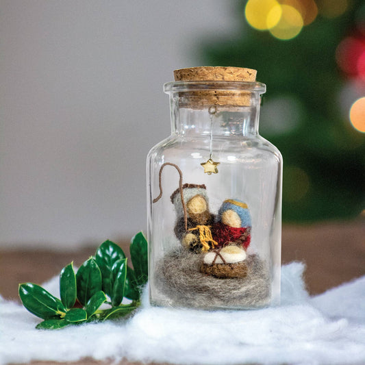 A clear glass bottle ornament with a cork stopper, containing a miniature nativity scene with felt figures, displayed on white faux fur with green leaves and a blurred Christmas tree in the background.
