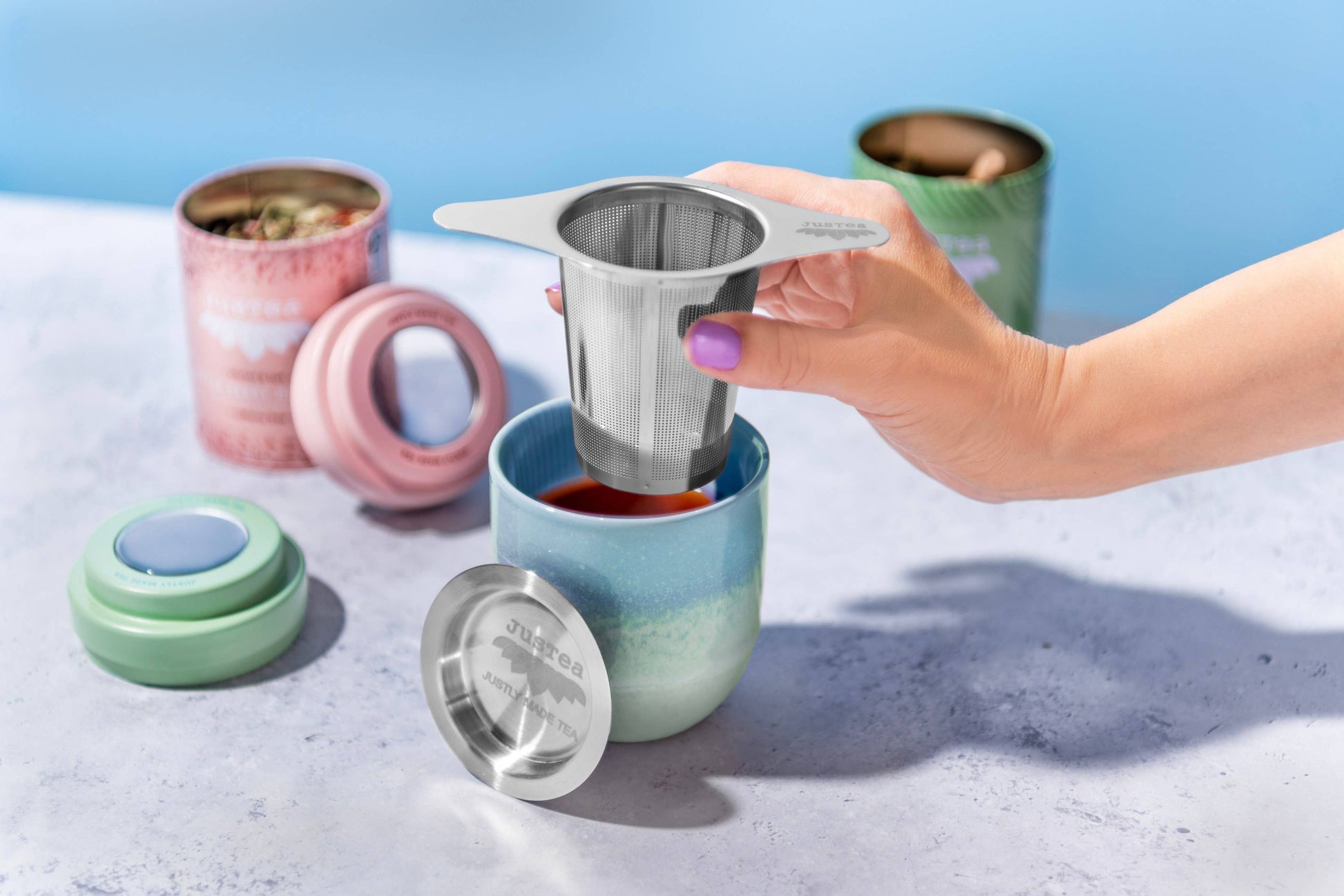 Person using a stainless steel tea infuser in a colorful mug with various tea accessories on a light blue background.
