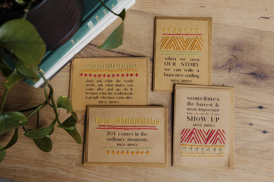 Four brown recycled paper cards with motivational quotes from Brene Brown styled on a wooden surface with a plant.