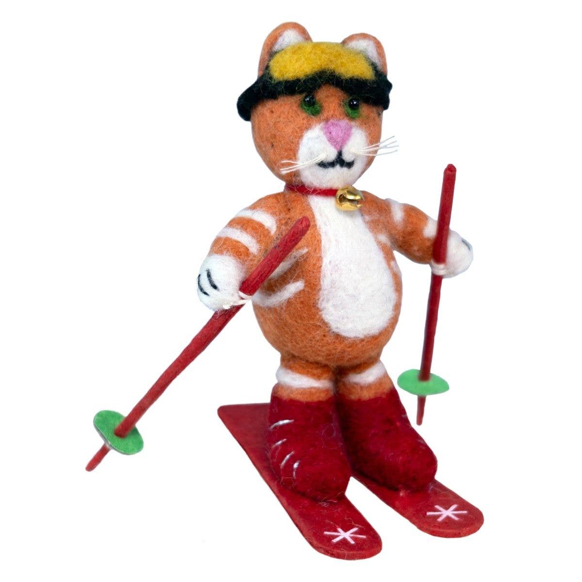 A felt ornament of an orange and white cat dressed as a skier, wearing a yellow and black hat, red boots, and holding red ski poles with green tips on a plain white background.