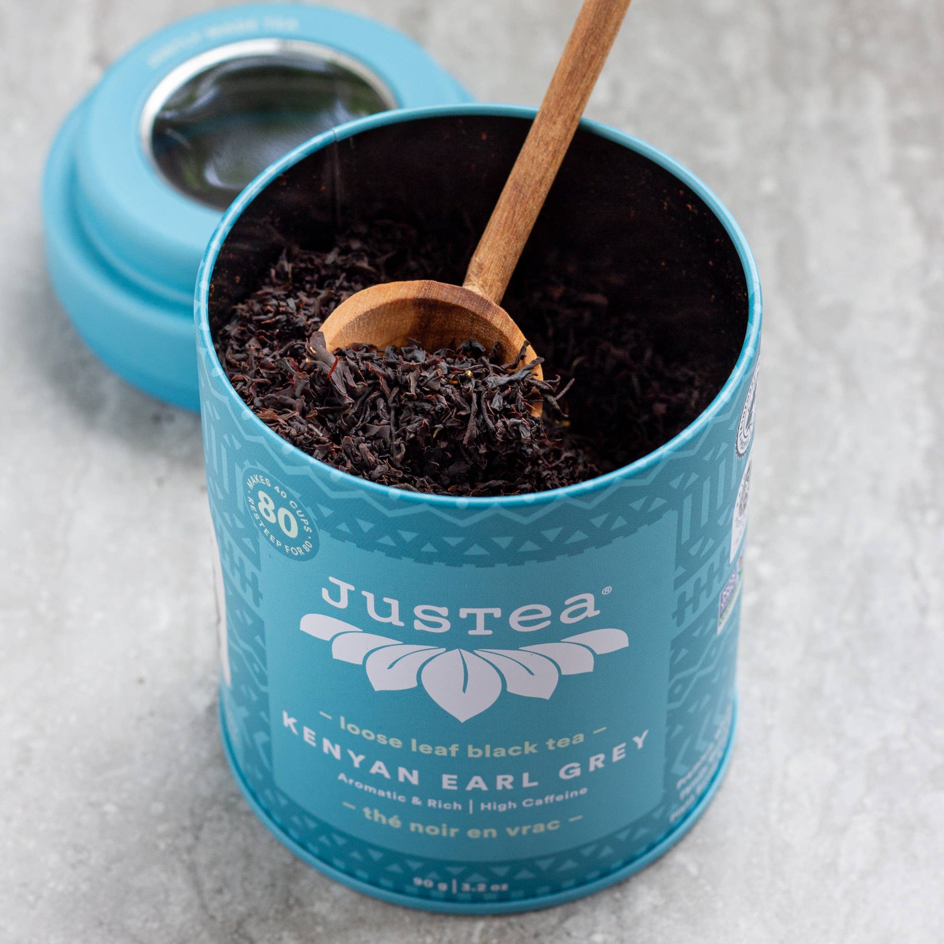 Teal canister of JusTea Kenyan Earl Grey black tea with a wooden scoop on a light gray background