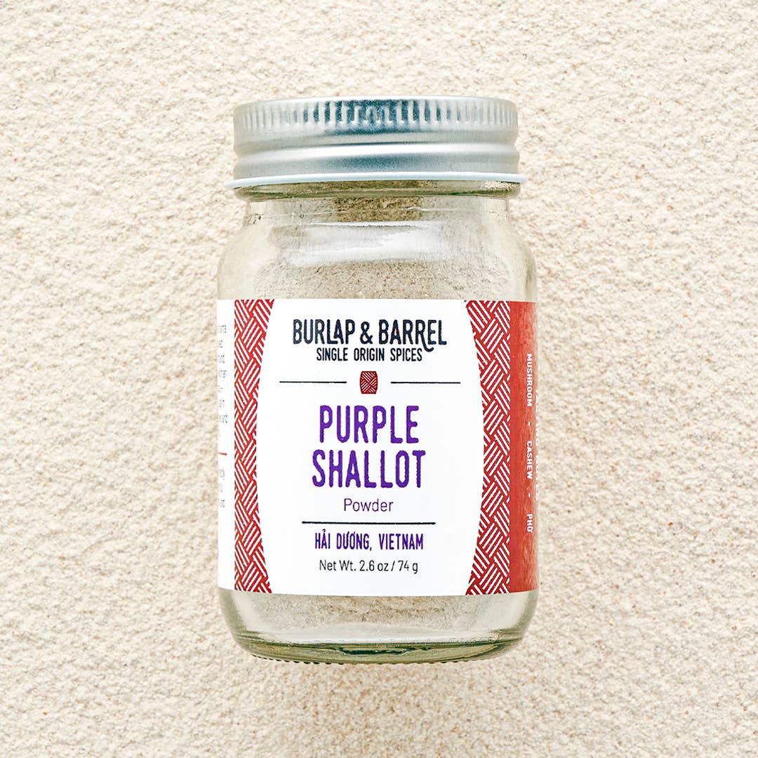 A glass jar of Burlap & Barrel Purple Shallot Powder, featuring a white label with purple text and red patterned side borders, sealed with a silver metal lid, set against a shallot powder backdrop.