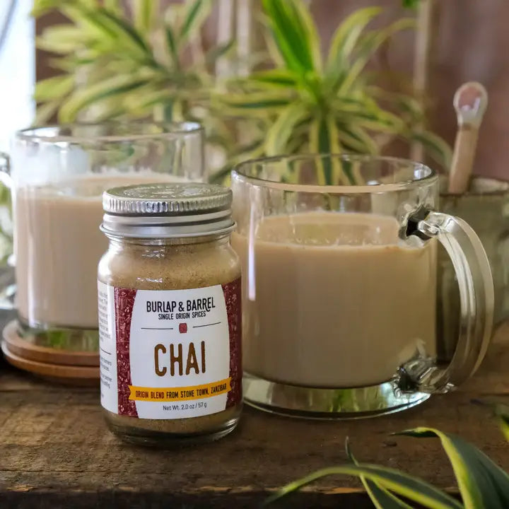 Jar of BURLAP & BARREL Chai spice mix with a glass mug of chai tea on a wooden surface.