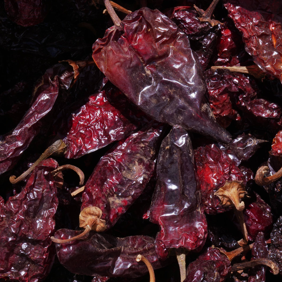A close-up pile of dried red chili peppers with wrinkled skin and visible stems, filling the frame against a very dark background.