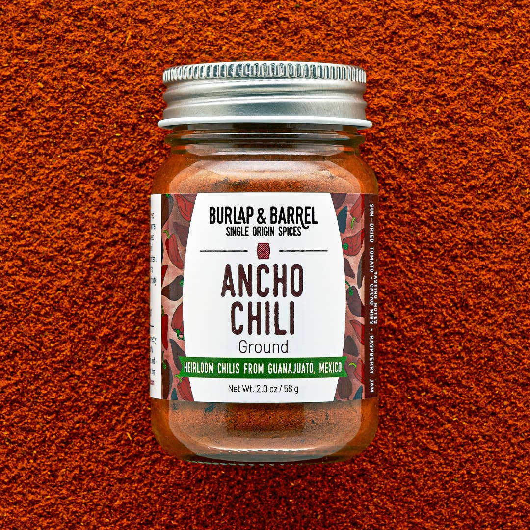 A jar of Burlap & Barrel Ancho Chili Ground spices, featuring a white label with red and green accents and a silver metal lid, set against a textured orange-red background of chili powder.