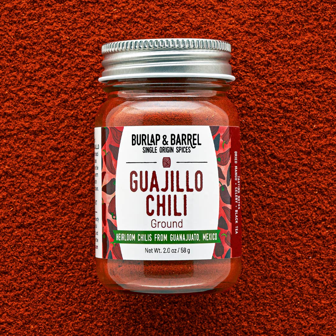 A jar of Burlap & Barrel Guajillo Chili Ground spice, featuring a white label with red and green accents and a silver metal lid, set against a textured red-orange background.