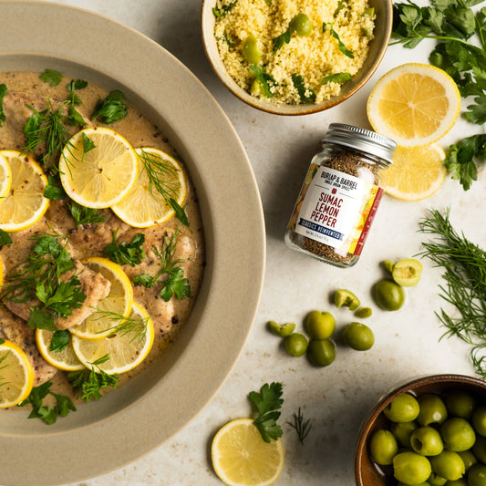 Dish with lemon slices and herbs, jar of sumac lemon pepper, and bowls of couscous and olives on a light surface.