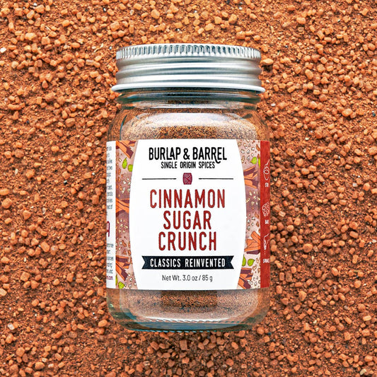 A glass jar of Burlap & Barrel Cinnamon Sugar Crunch spices, featuring a white label with red and black text, a silver metal lid, and a background of reddish-brown ground spices.