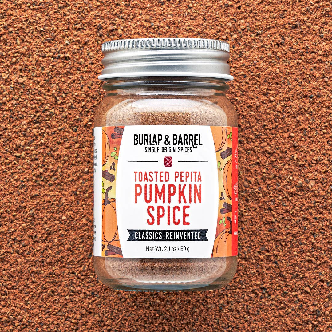 A jar of Burlap & Barrel Toasted Pepita Pumpkin Spice centered on a textured brown background. The clear glass jar shows the warm brown spice blend inside, topped with a silver metal lid and featuring a white label with orange and red illustrations.