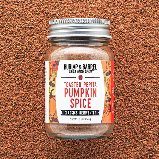 A jar of Burlap & Barrel Toasted Pepita Pumpkin Spice centered on a textured brown background. The clear glass jar shows the warm brown spice blend inside, topped with a silver metal lid and featuring a white label with orange and red illustrations.