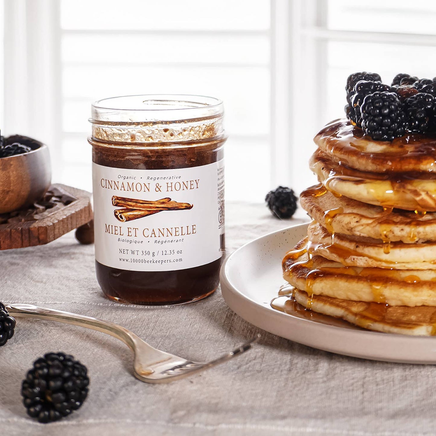 Jar of cinnamon honey next to a stack of pancakes with blackberries on a table.