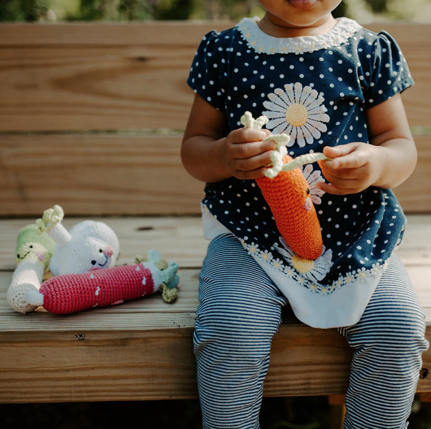 Child sitting on a wooden bench holding a crochet carrot toy, with other toys nearby.