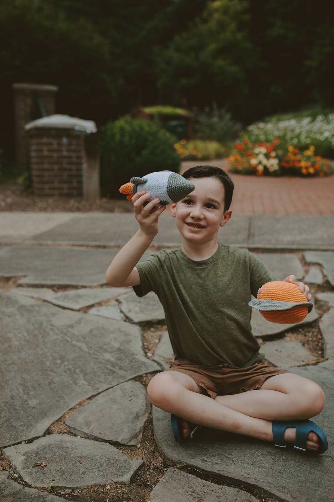 Child sitting on stone steps holding a toy rocket and planet, with a garden in the background