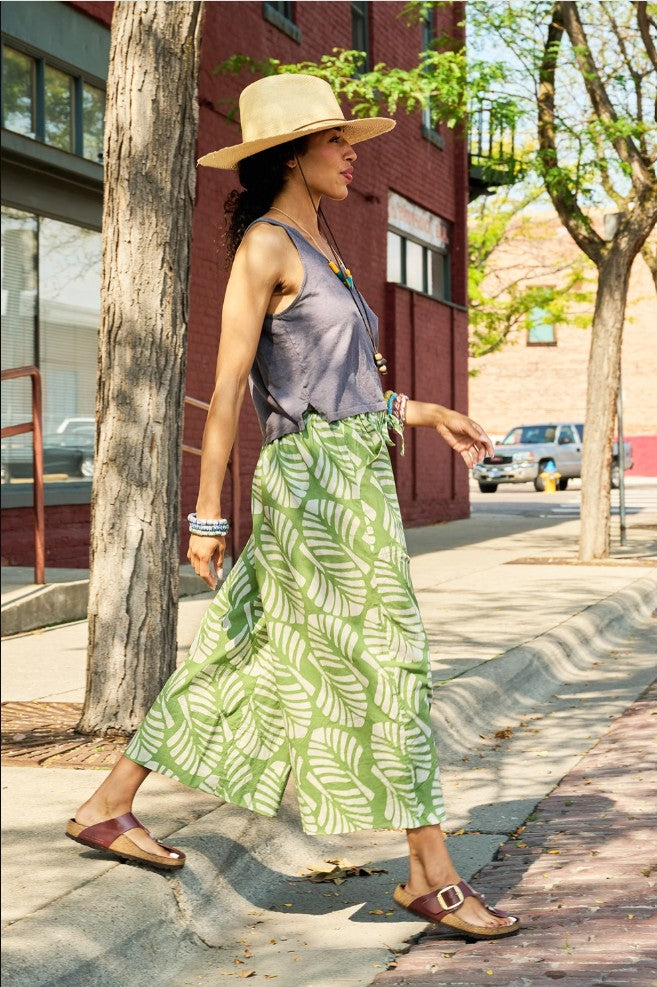 Woman walking on a sidewalk wearing a straw hat, gray top, and green leaf-patterned pants.