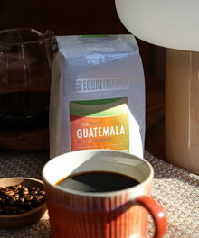 Equal Exchange Guatemala coffee bag on a burlap sack with a cup of brewed coffee and a bowl of coffee beans