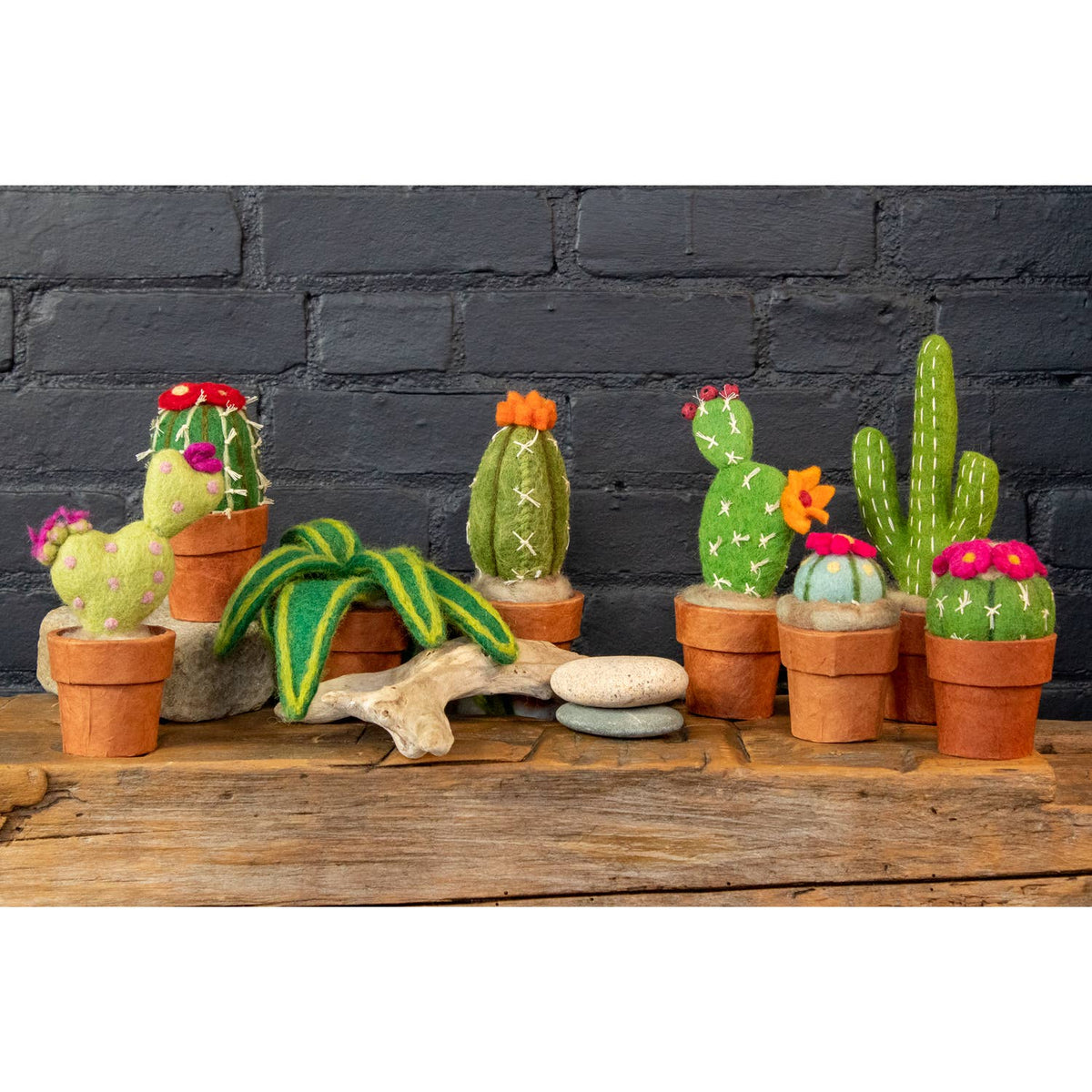 A set of felted wool cactus and succulent plants in terracotta pots, arranged on a rustic wooden surface against a dark gray brick wall background.