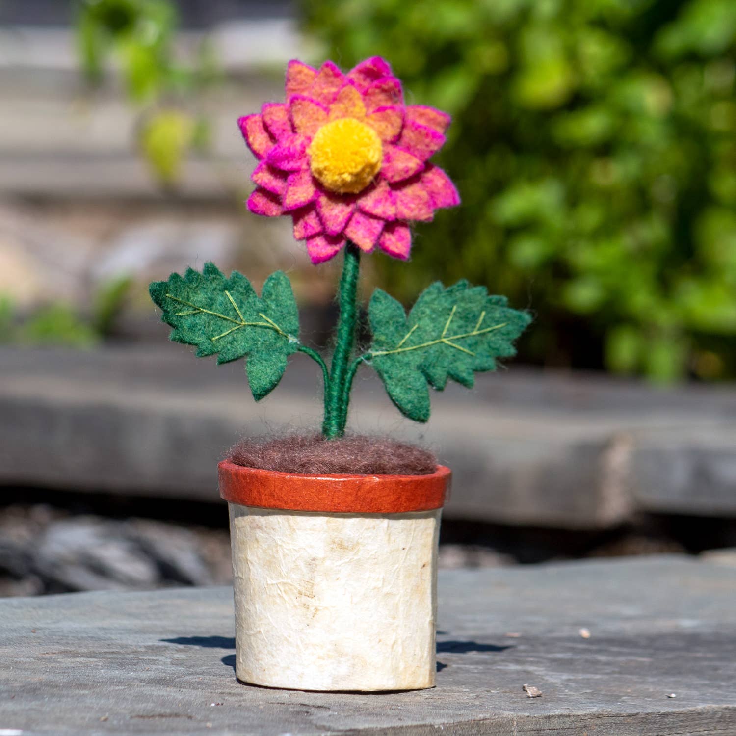 Felted flower in a pot on a wooden surface with blurred greenery in the background