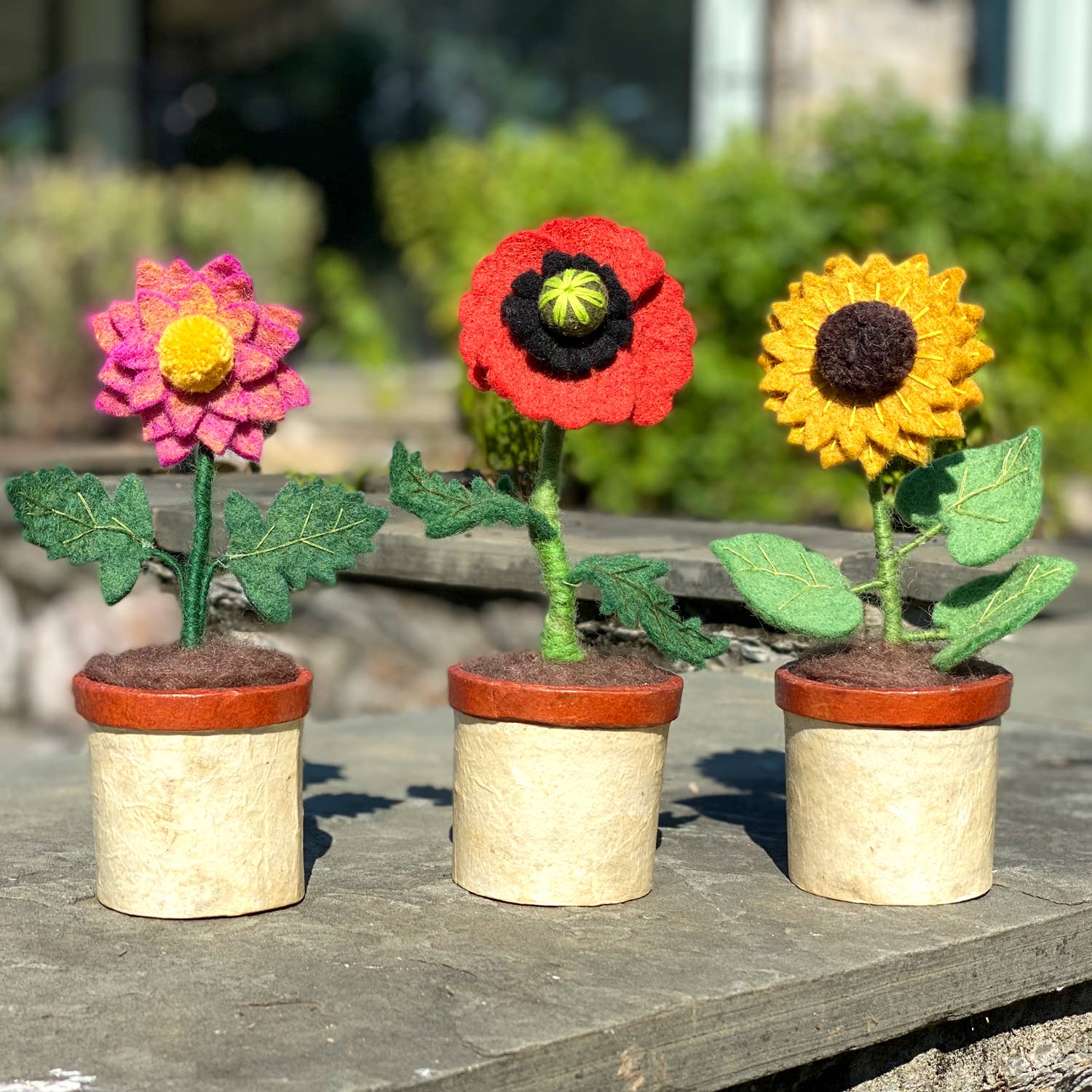 Three colorful felted flowers in paper pots on a wooden surface.