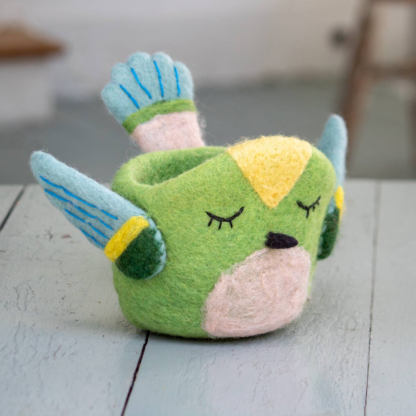 Felted green hummingbird with light blue and yellow details on a gray wooden surface. 