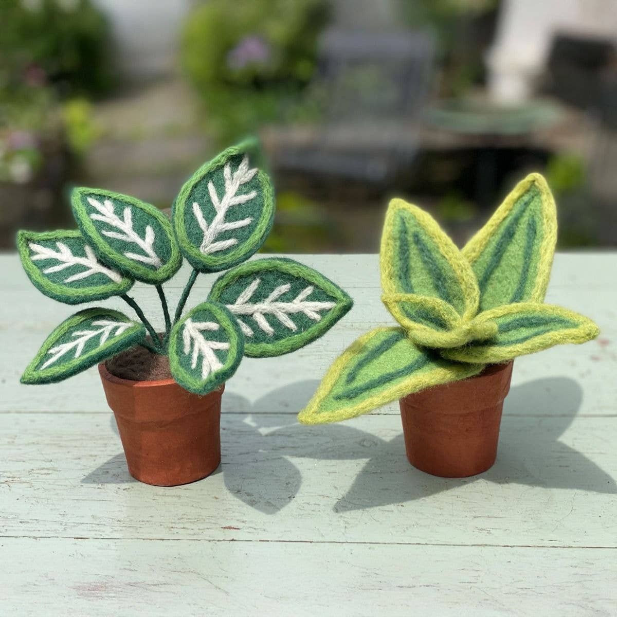 Two felt plants in terracotta-colored pots on a wooden surface with a blurred garden background.