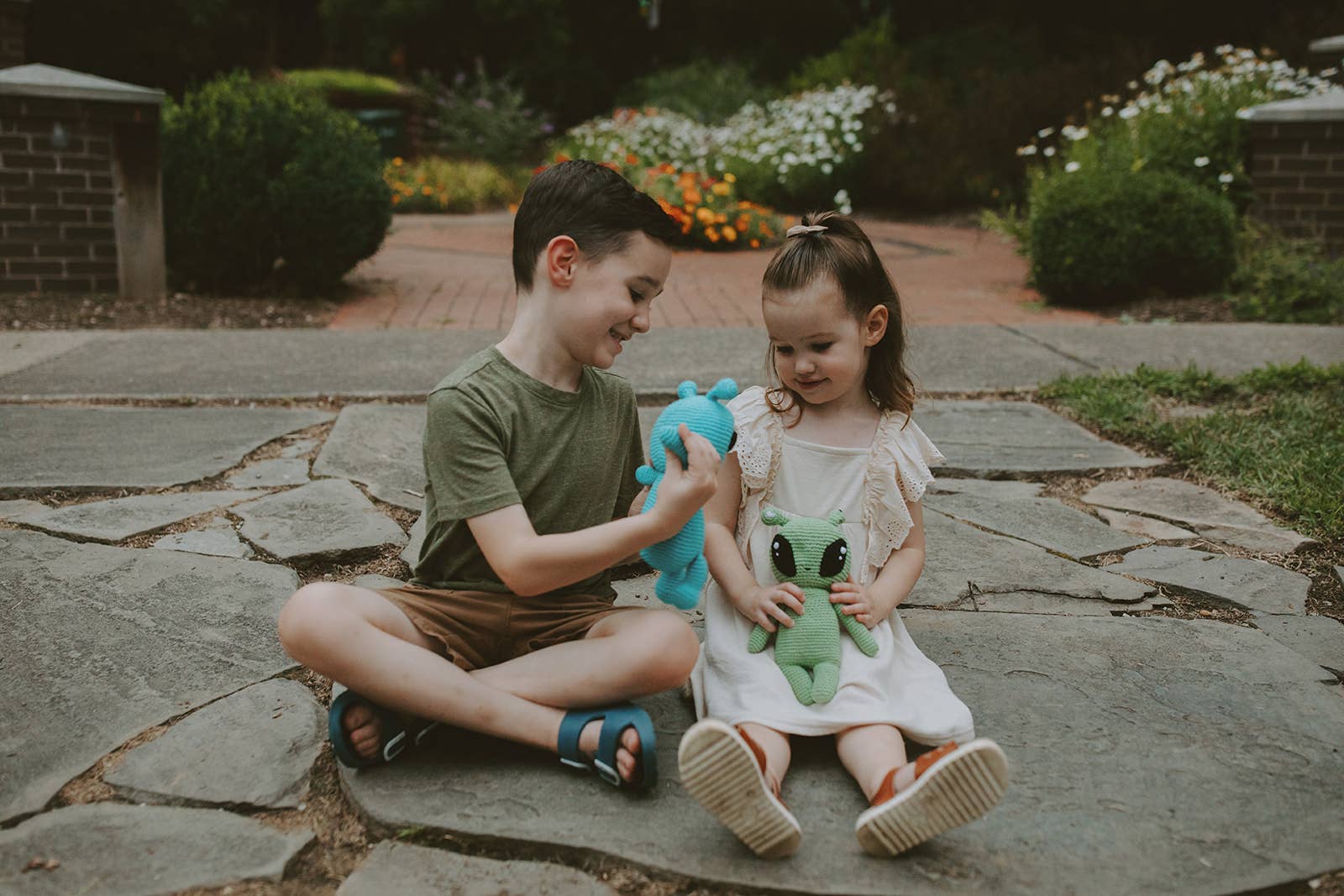 Two children sitting on a stone path outdoors, holding stuffed animals.