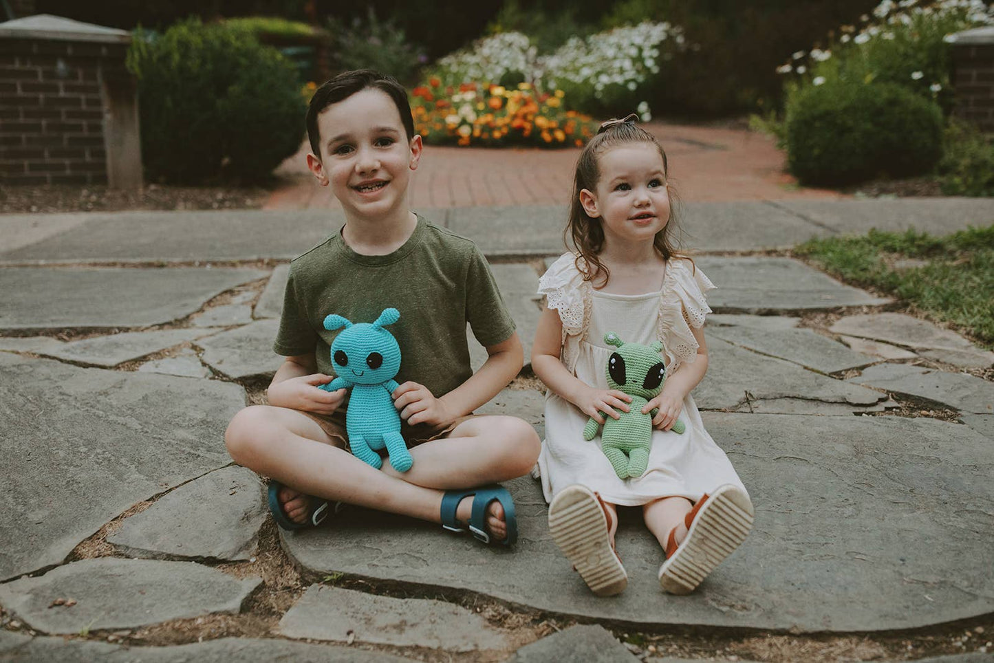 Two children sitting on a stone path holding stuffed aliens.