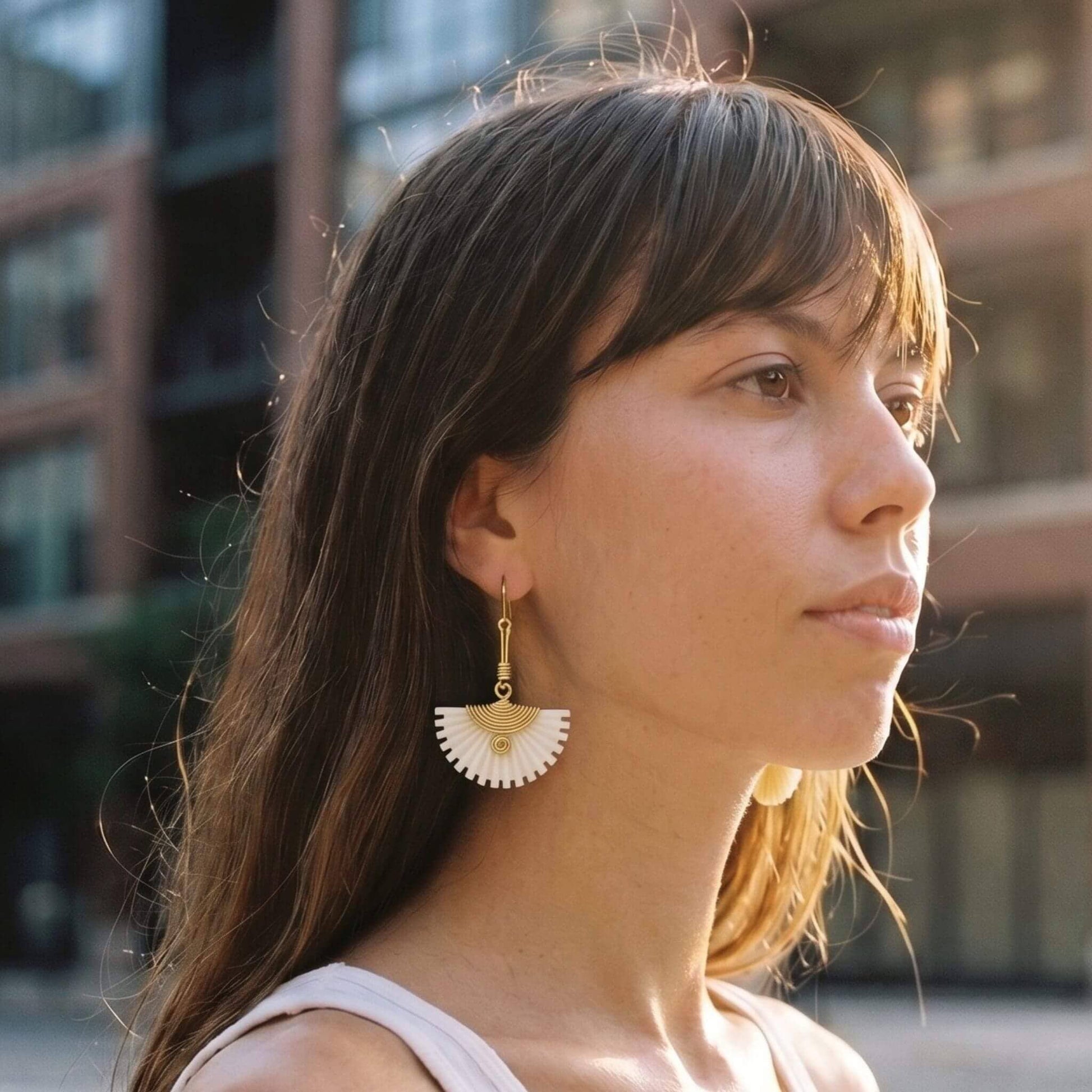 A woman in profile wearing a pair of gold and white fan-shaped dangle earrings, photographed outdoors with a blurred urban background.