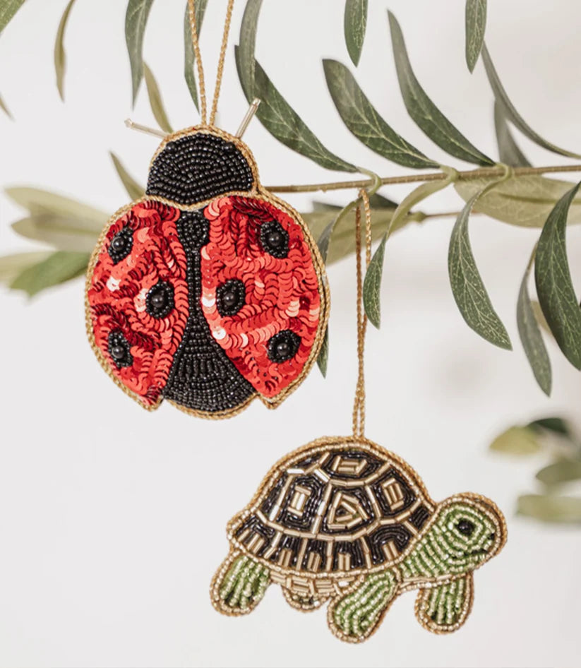 Green beaded turtle ornament and red beaded ladybug ornament hanging from leafy branches.