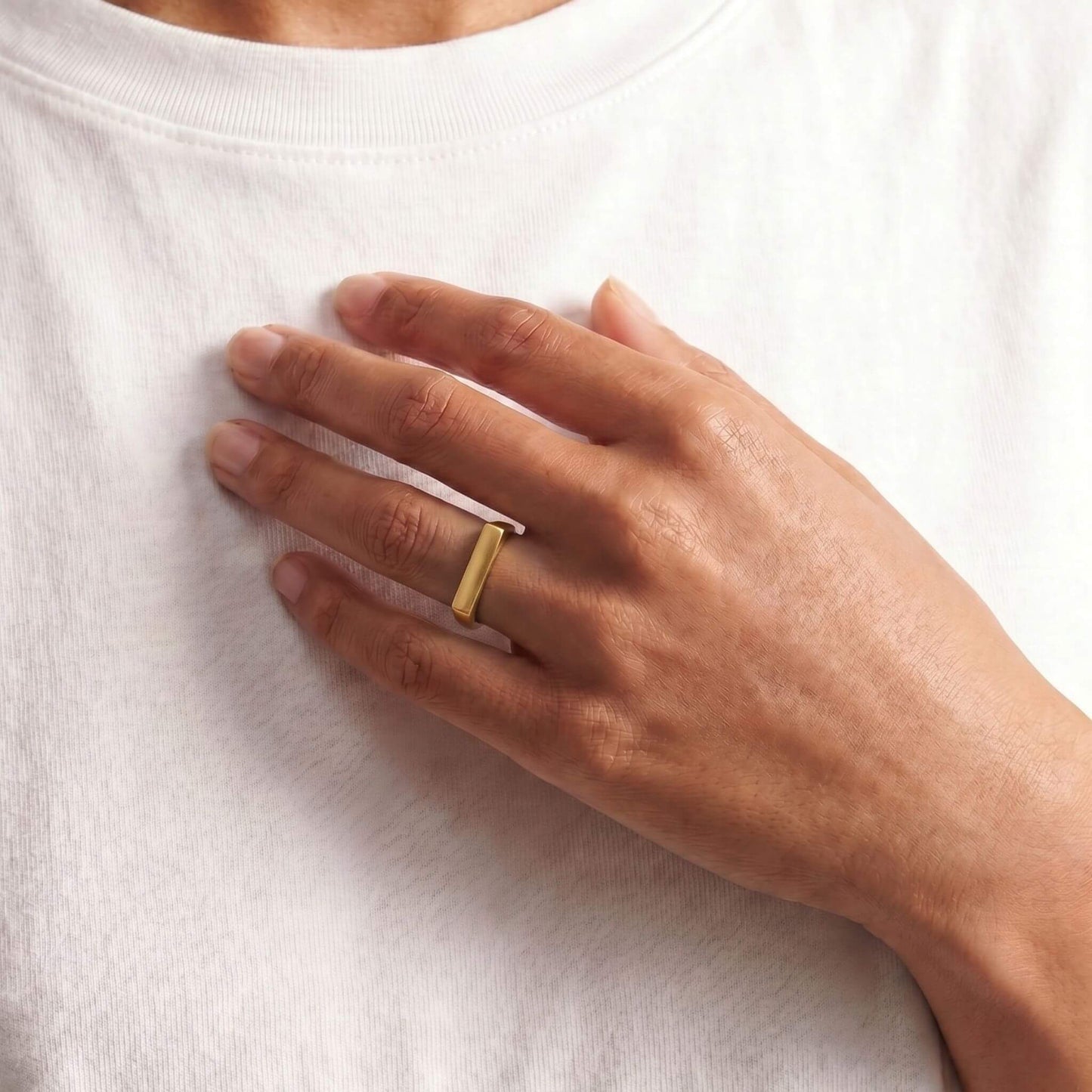 A close-up of a person's hand resting on their white t-shirt, wearing a simple gold band ring on one finger.
