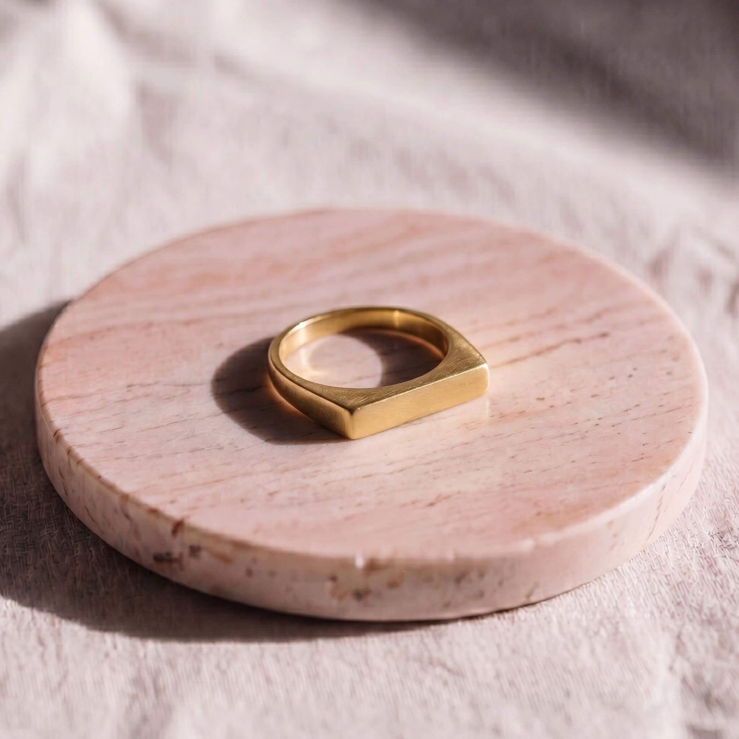 A gold ring with a wide, geometric square-shaped band, displayed on a round pink marble coaster over a textured beige fabric background.