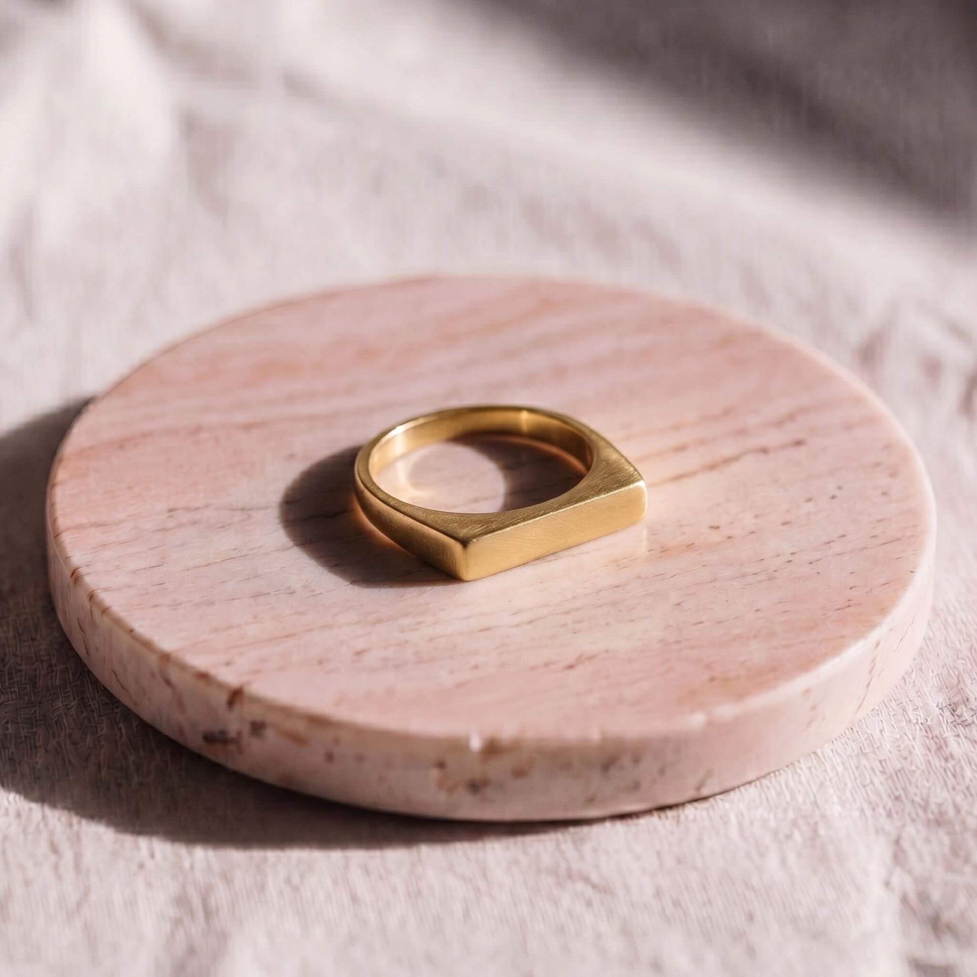 A gold ring with a wide, geometric square-shaped band, displayed on a round pink marble coaster over a textured beige fabric background.