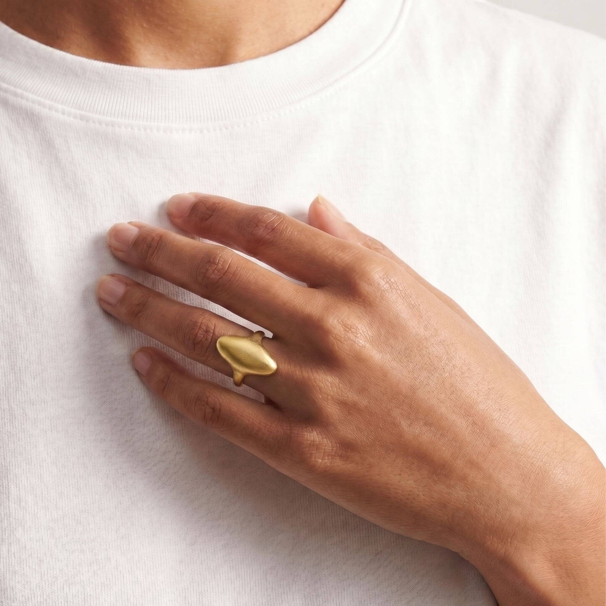 A close-up of a person's hand wearing a minimalist, organic-shaped gold ring on a white crew-neck t-shirt.