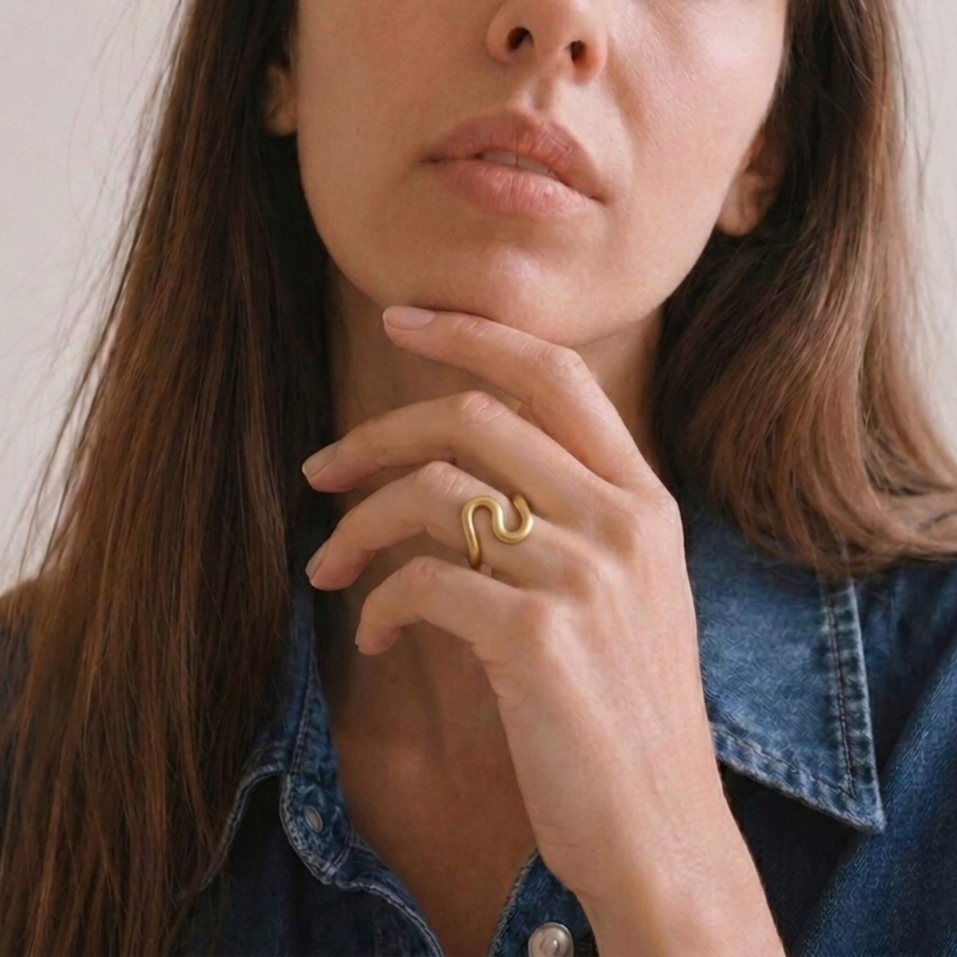 A close-up lifestyle shot of a woman wearing a gold ring with a wavy, snake-like design on her finger, paired with a blue denim shirt.