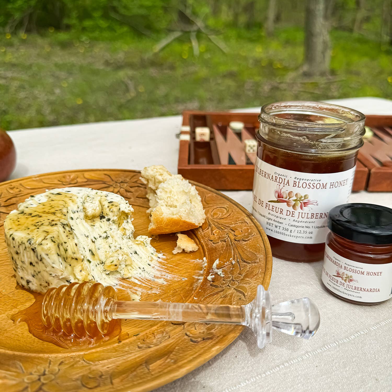 Wooden plate with cheese, bread, and honey, surrounded by jars of honey on a natural outdoor setting.