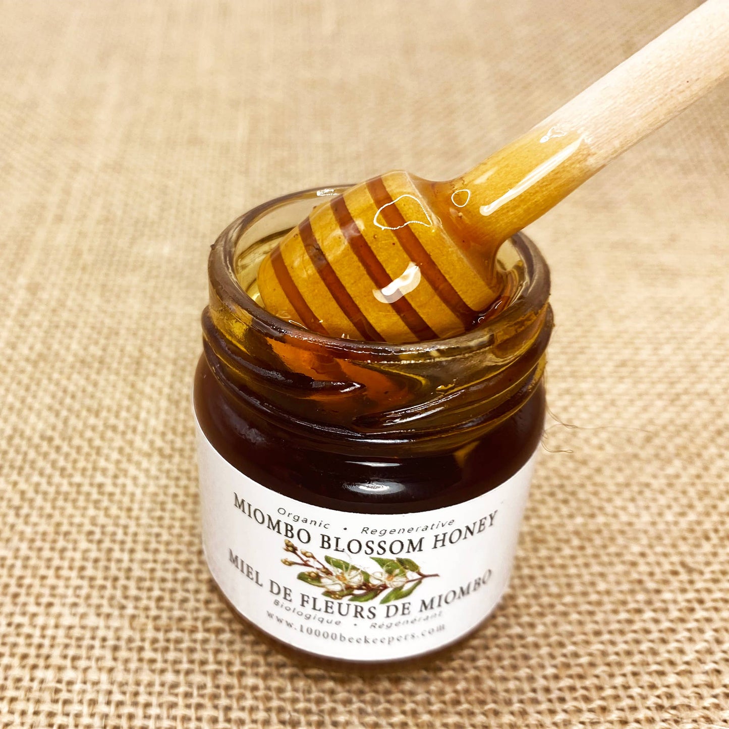 Jar of Miombo Blossom Honey with a honey dipper on a textured fabric background