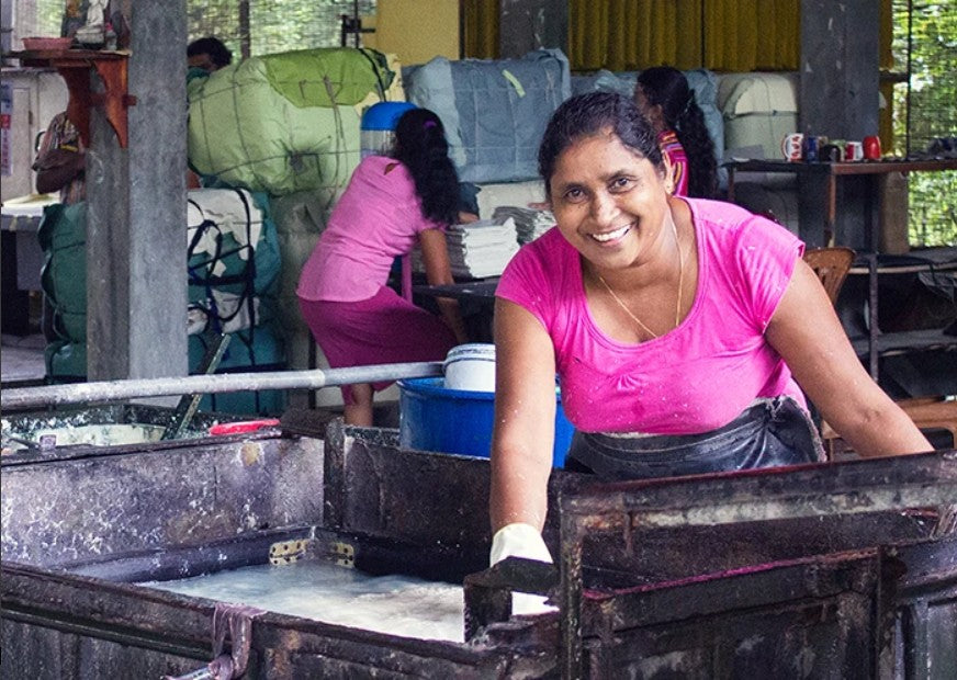 A smiling woman in a bright pink shirt leans over a large papermaking vat in a workshop, with other workers and colorful sacks visible in the background.