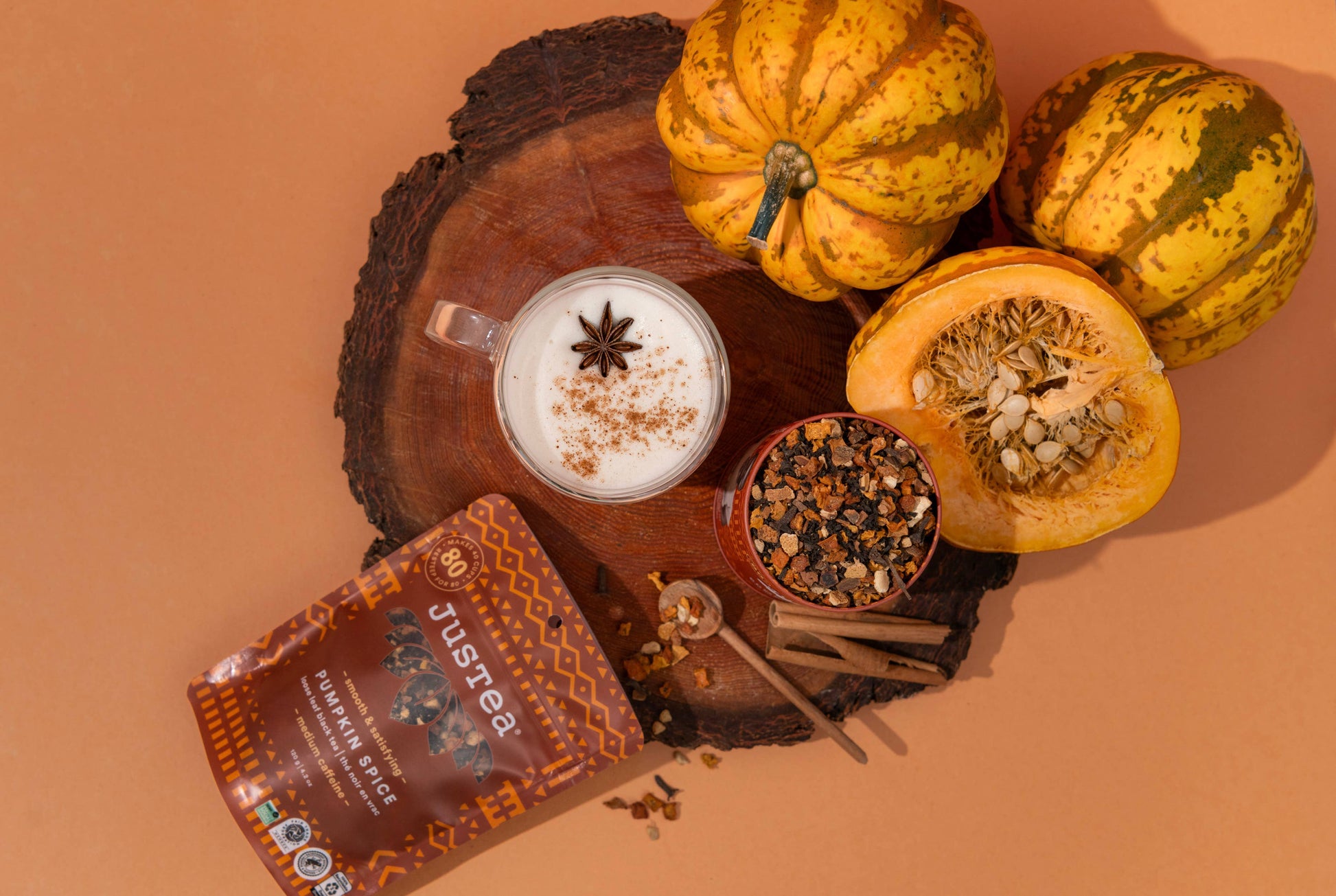 A styled flat-lay of Justtea Pumpkin Spice mix on a warm peach background, featuring a pouch of the product, a prepared iced latte with star anise, a small jar of the spice blend, cinnamon sticks, and three decorative pumpkins on a rustic wood slice.