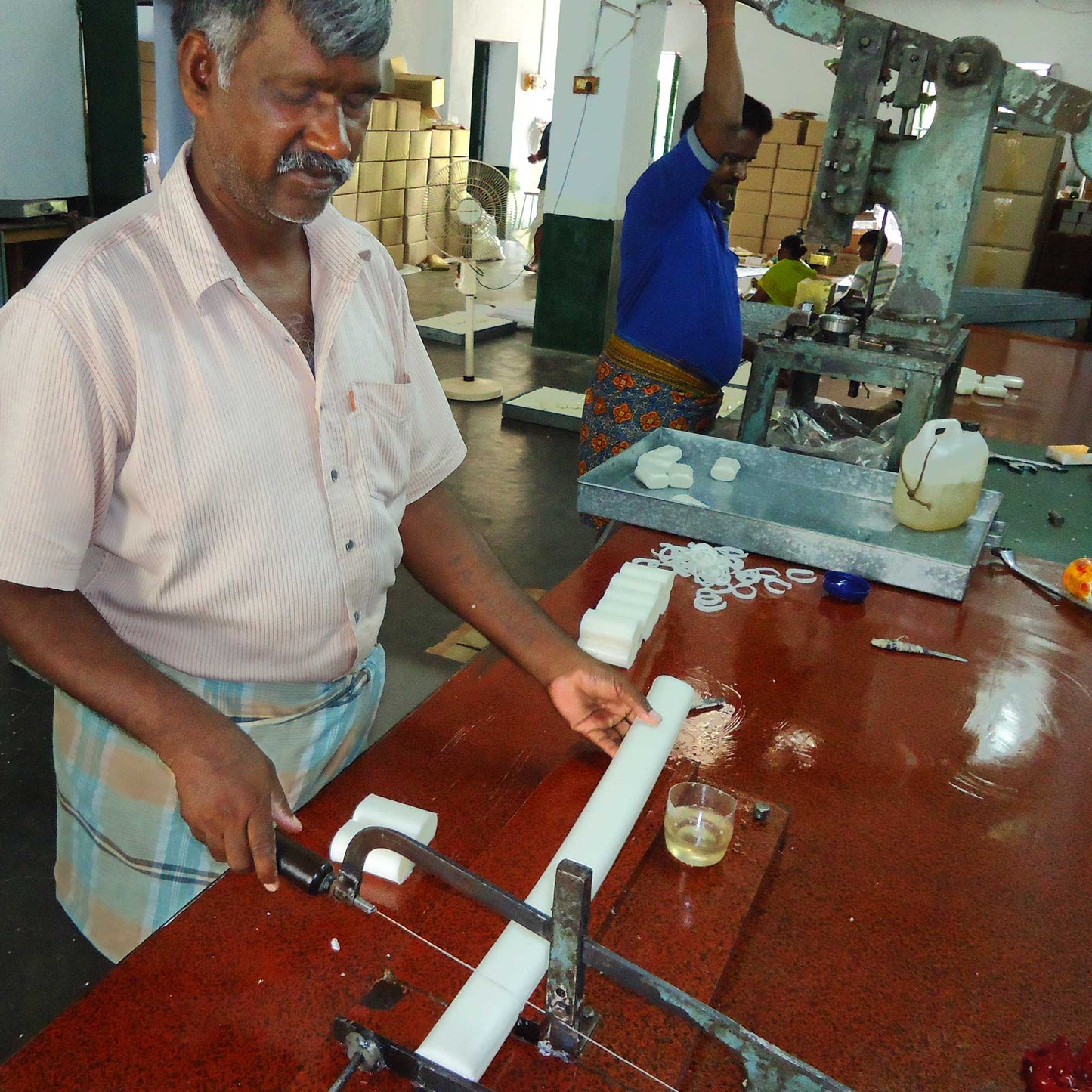 Two workers show off the process of cutting the bars of soap to size and pressing in the name "Palam" onto the finished bar of soap.