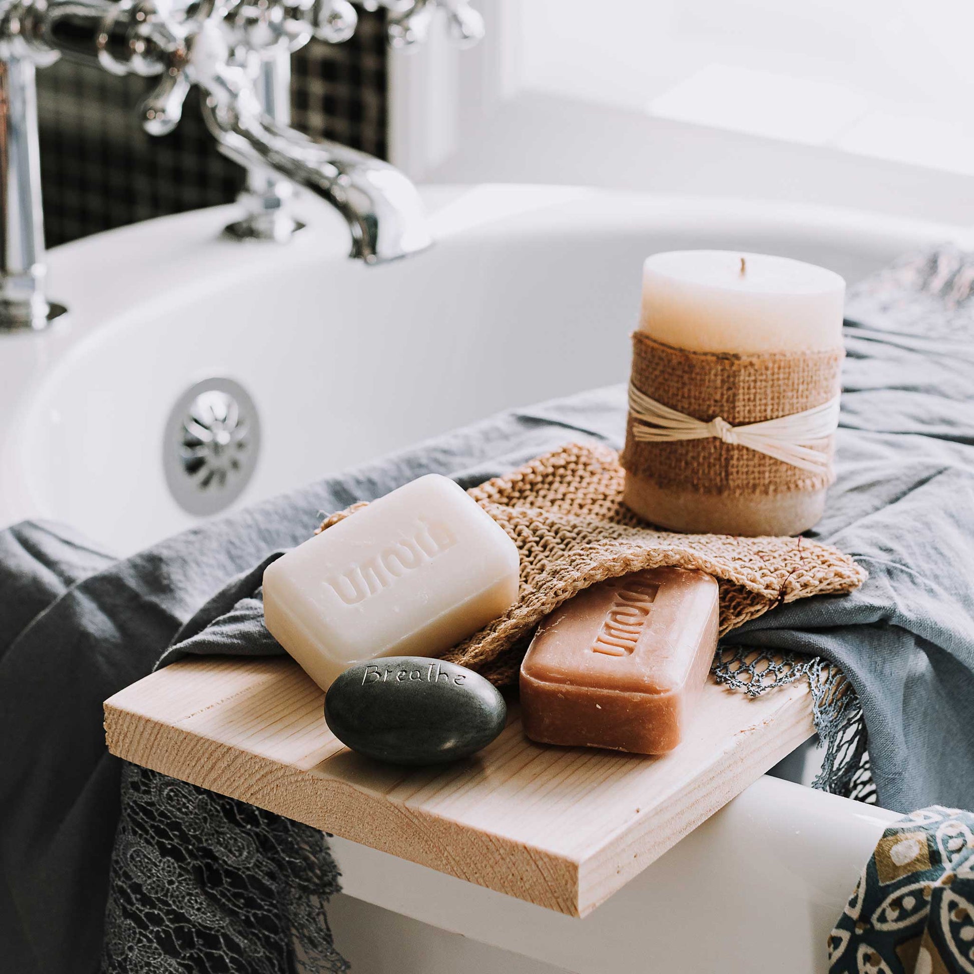 Two bars of soap, one white and one brown, are styled with a candle and a throw on the side of a white bathtub.