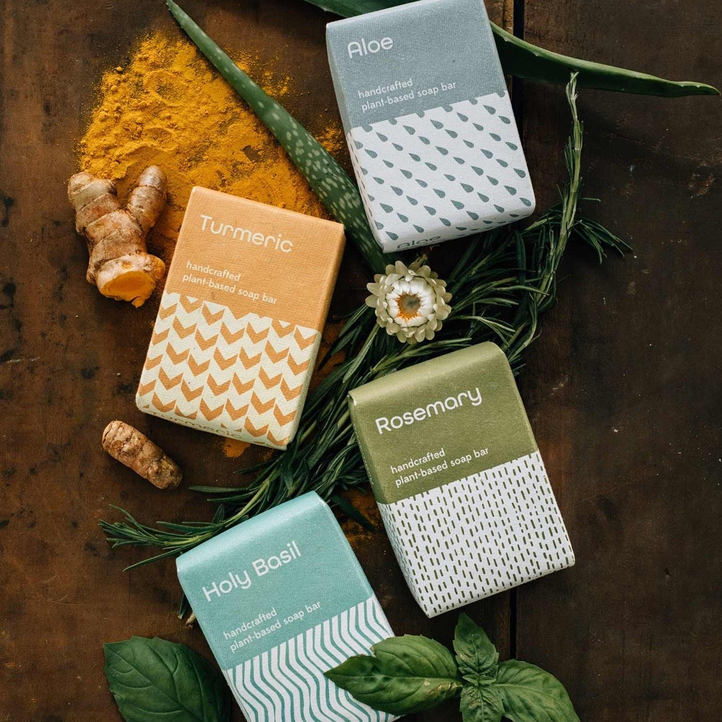 Four different bars of soap in their colored packaging are styled together on a wooded table top and surrounded by the ingredients that make them.