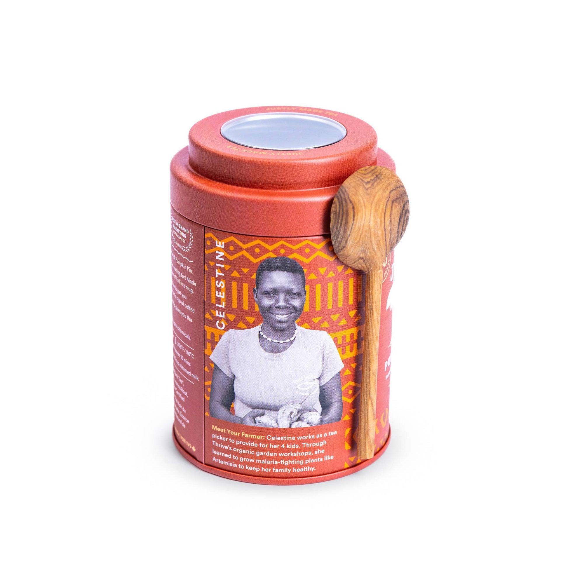 A cylindrical tea tin with a terracotta red lid and base, featuring an orange and black geometric patterned label with a portrait of a woman. A wooden spoon rests on the side of the tin, and the product is photographed on a clean white background.