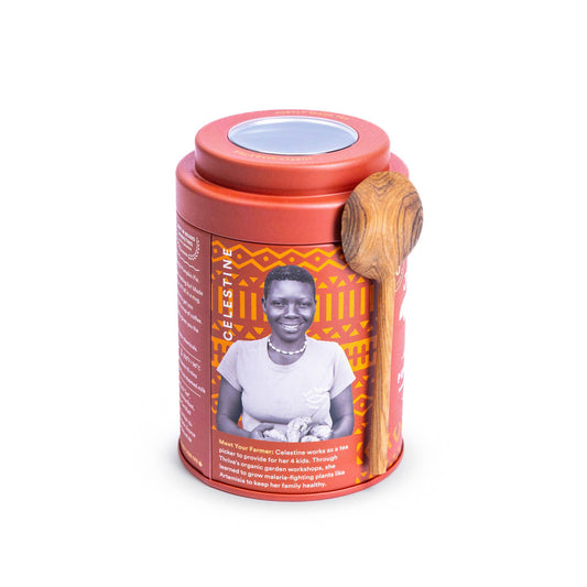 A cylindrical tea tin with a terracotta red lid and base, featuring an orange and black geometric patterned label with a portrait of a woman. A wooden spoon rests on the side of the tin, and the product is photographed on a clean white background.