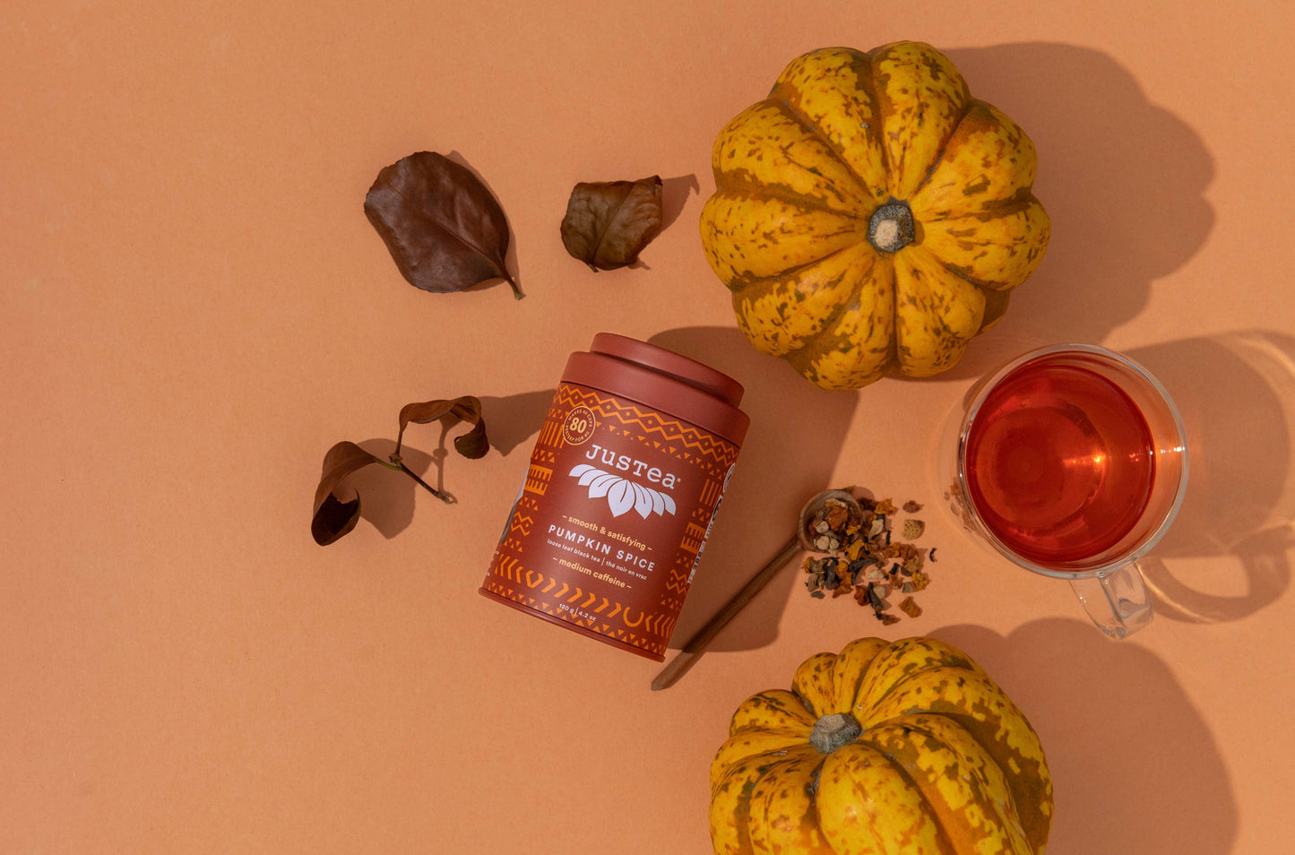 A styled flat-lay of Justtea Pumpkin Spice tea featuring a terracotta-colored tea tin, a glass mug filled with amber tea, loose tea leaves, cinnamon, dried leaves, and decorative pumpkins on a solid peach background.