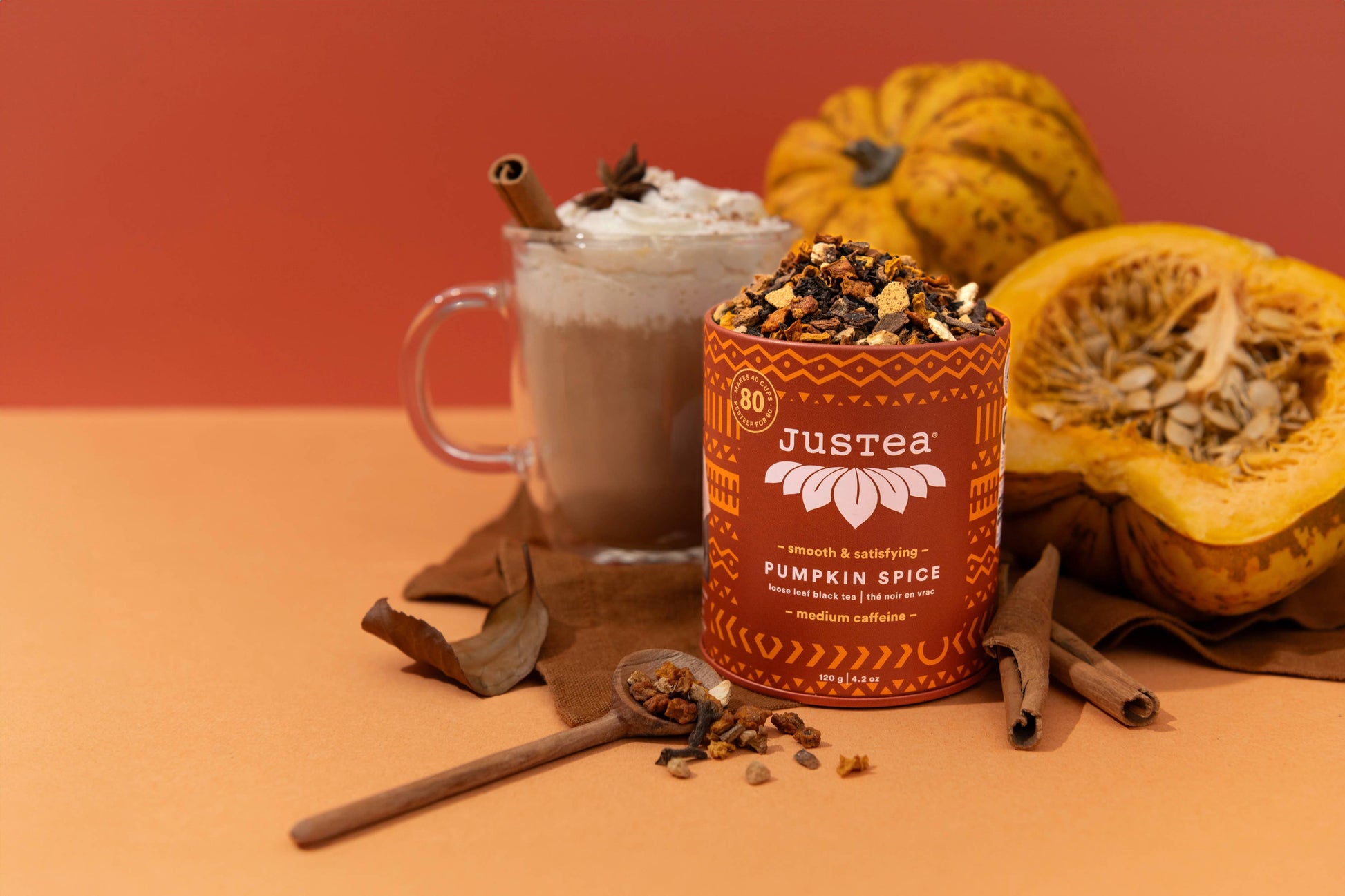 A styled product shot of Justtea Pumpkin Spice coffee tea in an orange cylindrical tin, set next to a whipped coffee drink with a cinnamon stick, with whole and halved pumpkins and cinnamon sticks on an orange surface against a solid orange background.