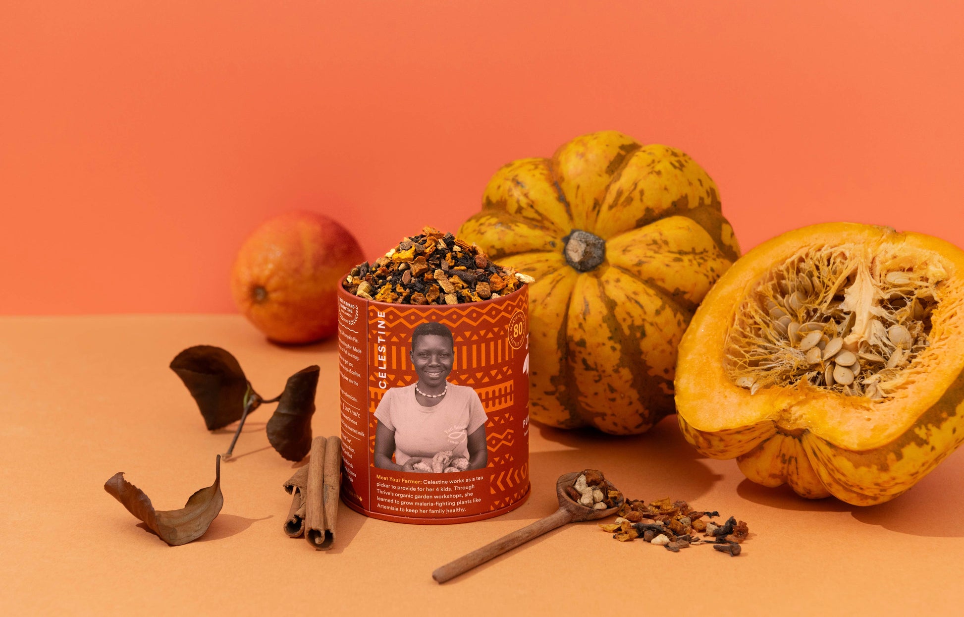 A tin of pumpkin spice tea with a portrait of a woman on the label, styled with whole and halved orange pumpkins, cinnamon sticks, dried leaves, and a wooden spoon on an orange surface against a solid orange background.