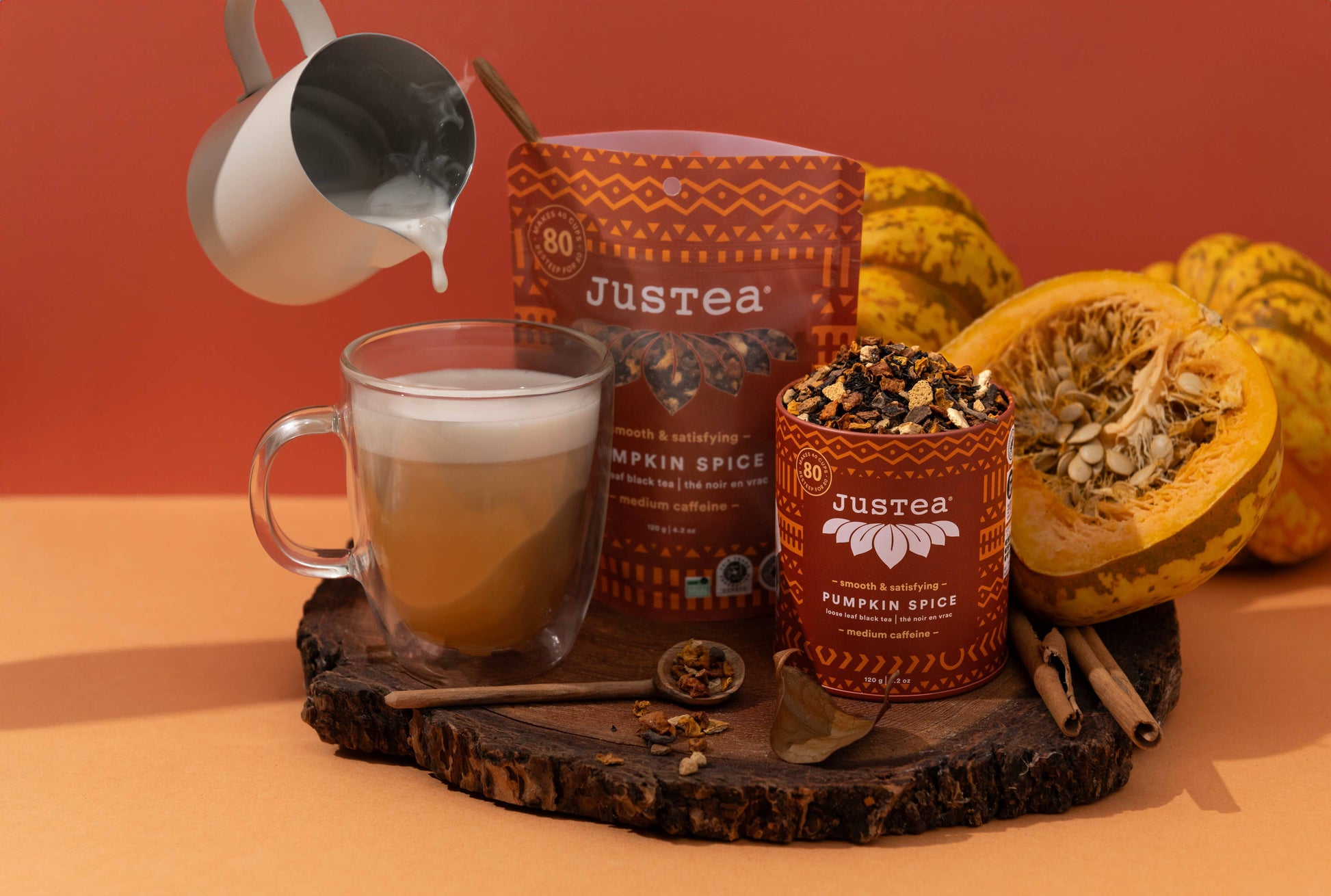 Justtea Pumpkin Spice coffee blend shown with a prepared latte being poured into a glass, a standing pouch, a canister of coffee, loose spice mix, cinnamon sticks, and pumpkin halves on a wooden slice against a warm orange background.