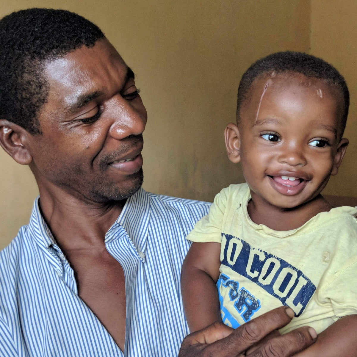 Man holding a child wearing a 'Too Cool' shirt against a beige background
